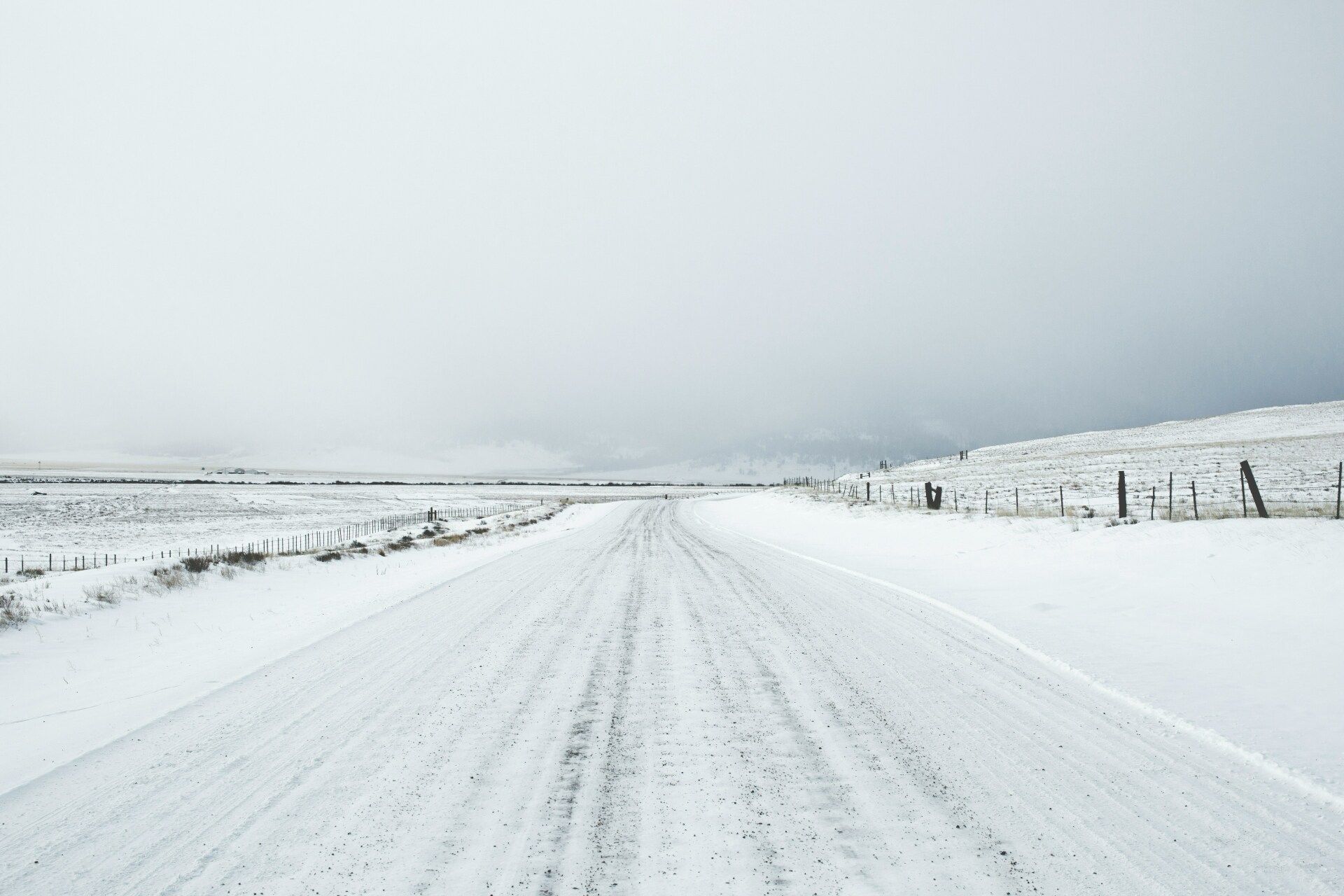 A snowy road in the middle of a field with a fence in the background.