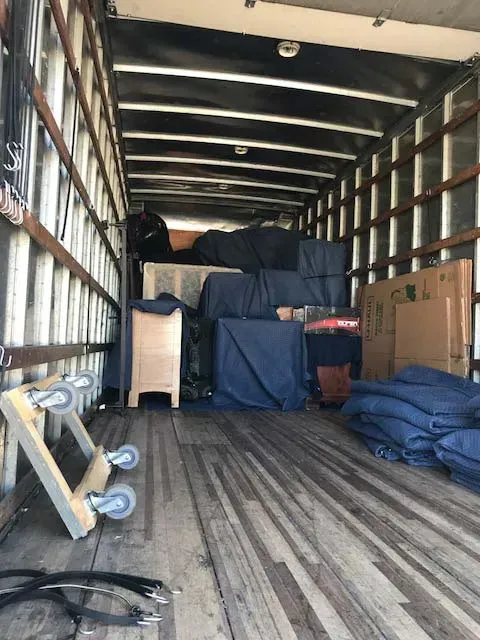 Two movers in red shirts and coveralls carrying boxes in a room with furniture wrapped in plastic.