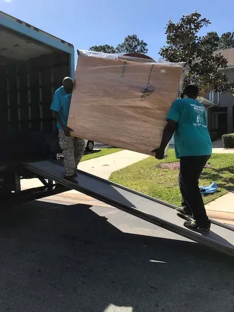 Two people in teal shirts move a large, wrapped rectangular item down a truck ramp.