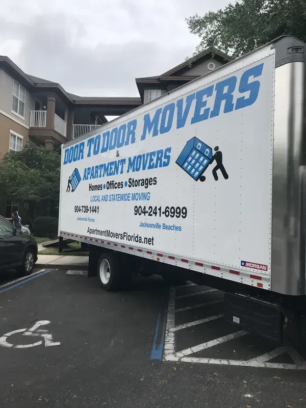 Moving truck parked in a handicapped spot in front of an apartment building. Door to Door Movers is printed on the side.