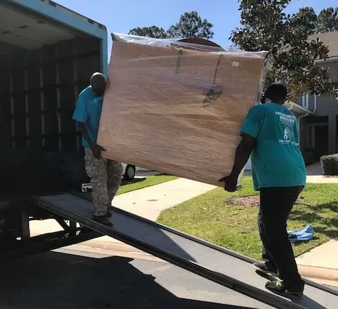 Two people carrying a large, wrapped object down a ramp from a truck. Two people carrying a large, wrapped object down a ramp from a truck.