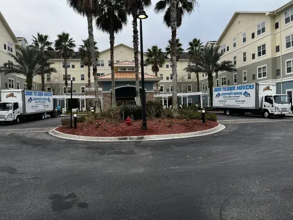 Two moving trucks parked outside a multi-story building entrance with palm trees.