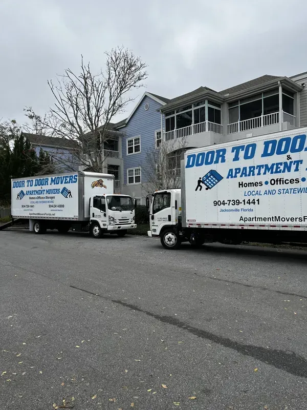 Two Door to Door Movers trucks parked on a street in front of an apartment building on a cloudy day.