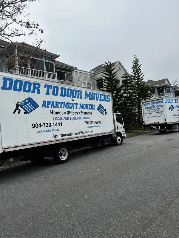 Two movers in blue overalls push a large wrapped object on a hand truck in a kitchen, boxes nearby.