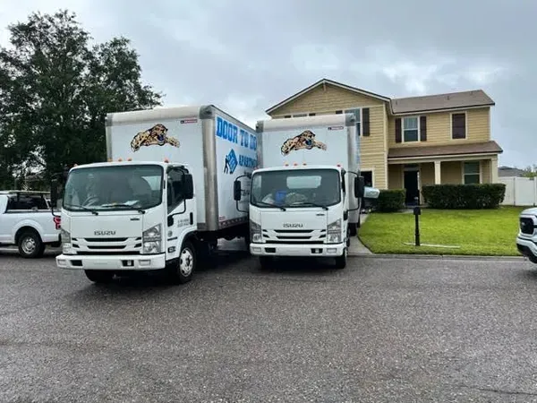 Two white moving trucks parked in front of a two-story house, preparing for a move on a cloudy day.