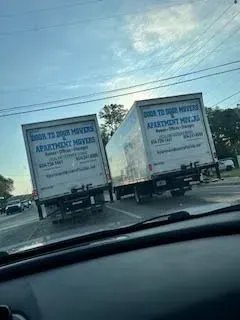 Two movers carrying furniture from a moving truck into a house with boxes on the ground.