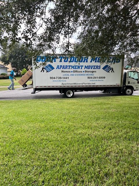 Moving truck parked on a grassy lawn with person carrying a box. Door to Door Movers is on the truck.