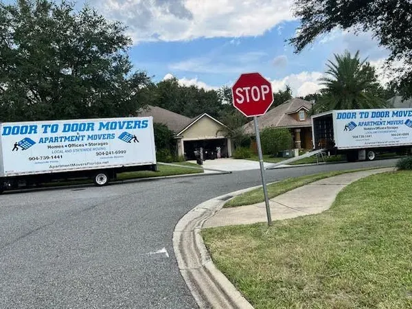 Two moving trucks labeled Door to Door Movers parked in front of a house, on a sunny day. Stop sign in foreground.
