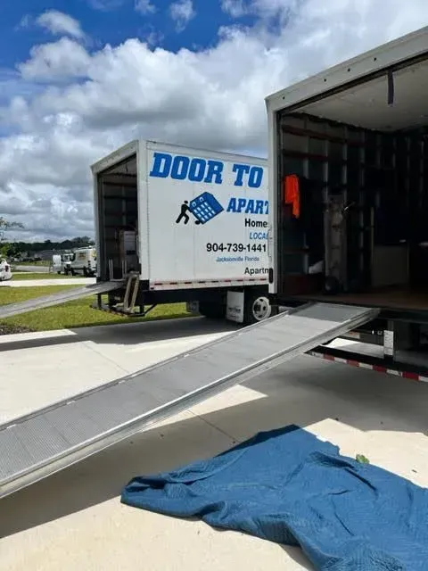 Two movers carrying a box into a sunlit living room with a sofa and stacked boxes.