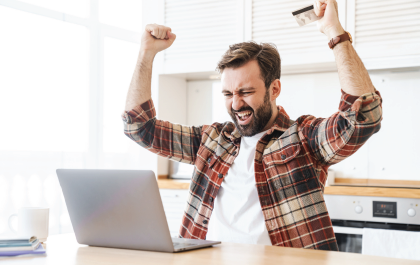 Man celebrating with raised fists and credit card near a laptop. Kitchen setting.