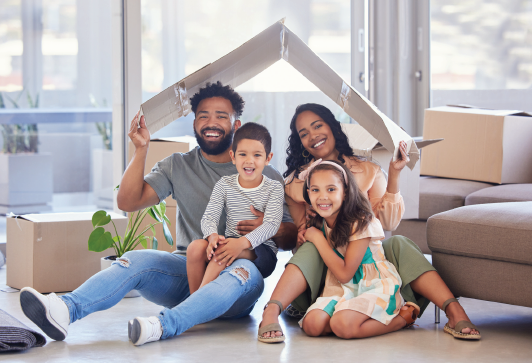 Family of four sitting under a cardboard roof, smiling in a room with moving boxes.
