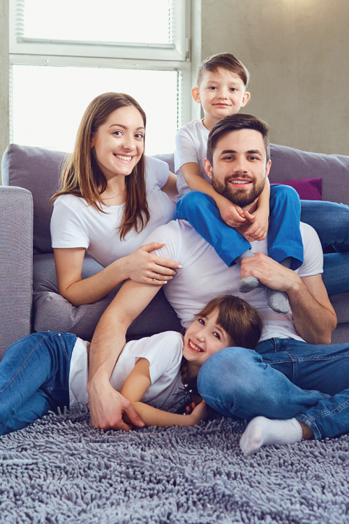 Family of four smiles, posing on the floor near a couch. Children on father's shoulders, mother sits near.