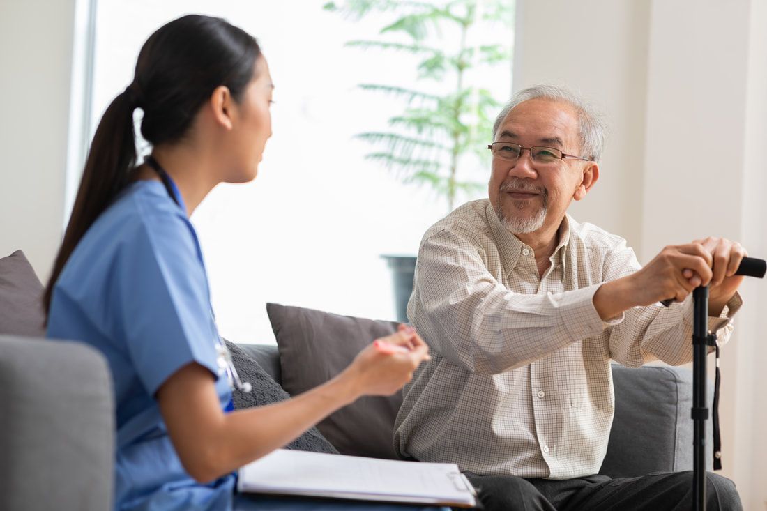 Caregiver talking with a person seated on a couch holding a cane. They are in a home setting.