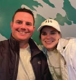 Smiling people in front of a turquoise wall. Man in dark jacket; woman in a white cap.
