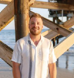 Man with a beard smiles under a wooden pier at the beach, wearing a striped shirt.