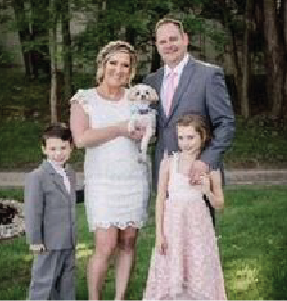 Family portrait: woman holding small dog, man in suit, children in formal wear, outdoor setting.