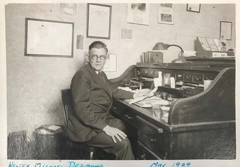 Man seated at desk in office, wearing glasses, suit. Papers and lamp on desk; framed documents on wall.