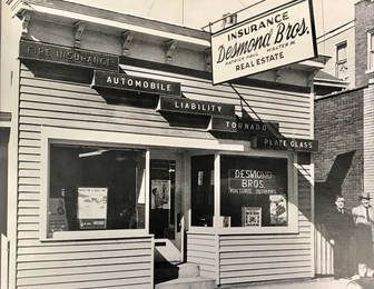 Black and white photo of Desmond Bros. Insurance and Real Estate office building with signs for various insurance types.