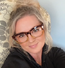 Woman with blonde hair in a bun wearing tortoise-shell glasses, smiling, resting on a floral cushion.