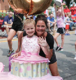 Woman and girl in pink dress pose with cake prop at an outdoor event, smiling.