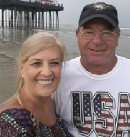 Couple smiling on a beach, pier in the background. Man in USA shirt and cap. Woman in patterned top.