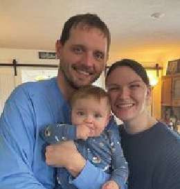 Family smiling indoors, man holding child. Blue shirts, warm lighting.