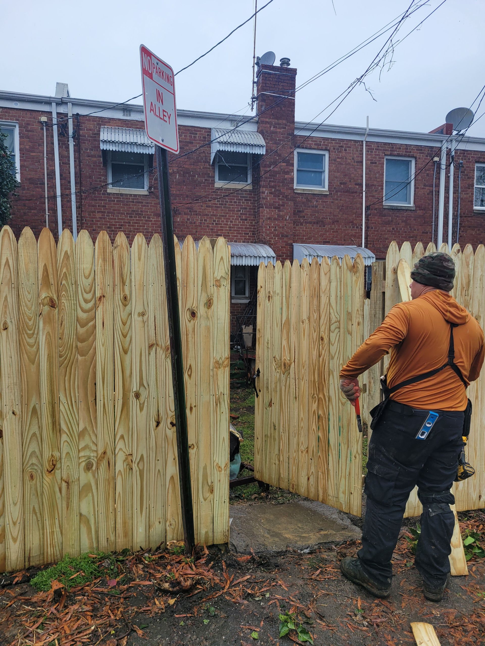 A man is standing next to a wooden fence in front of a brick building.