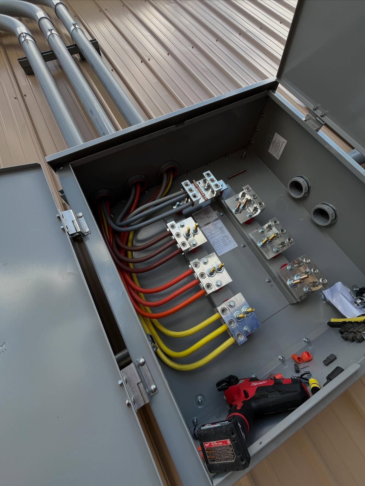 Open electrical box on a metal roof, containing wires, breakers, and conduit. A power drill rests inside.