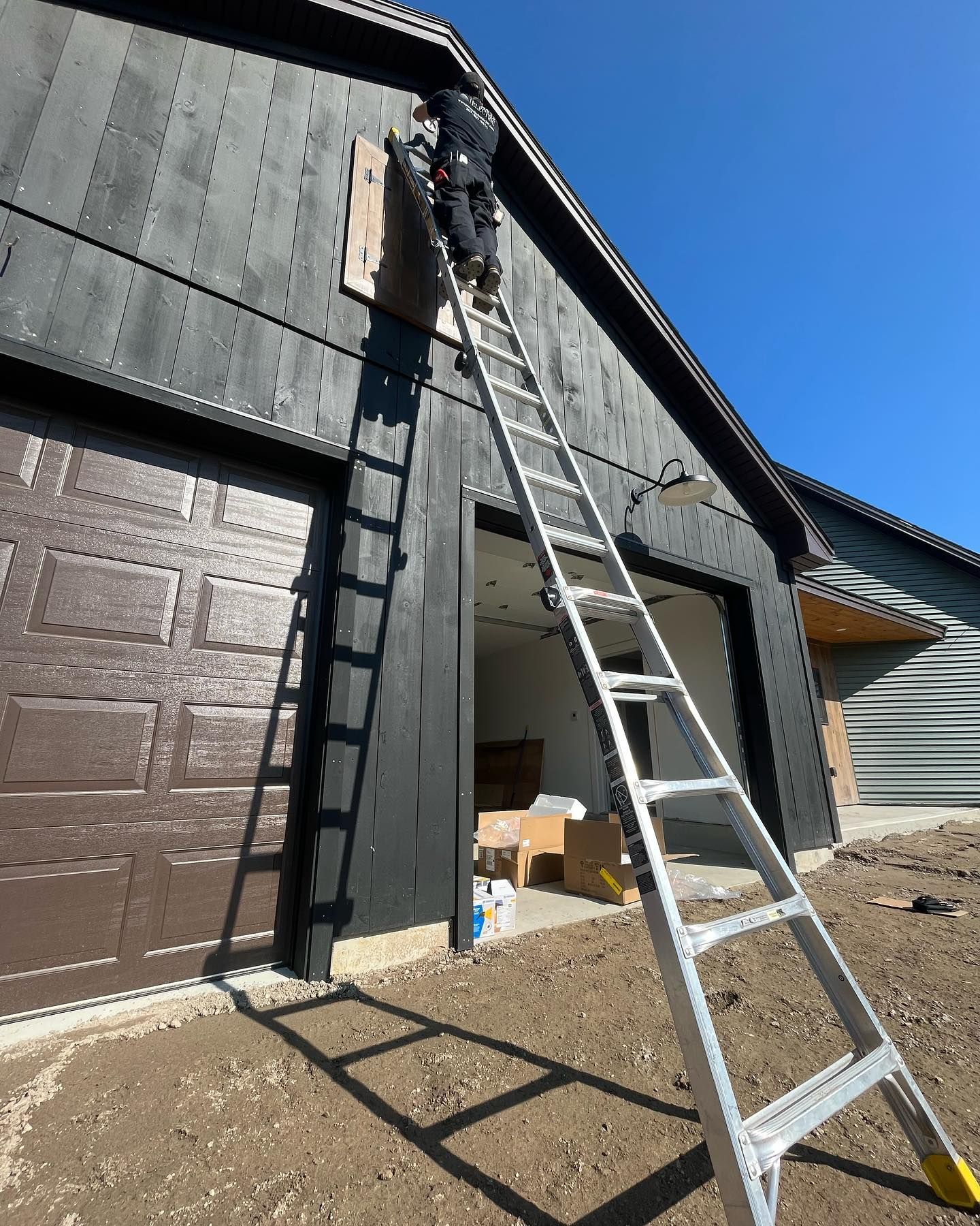 Person on a ladder working on dark exterior wall above a garage door. Sunny day.