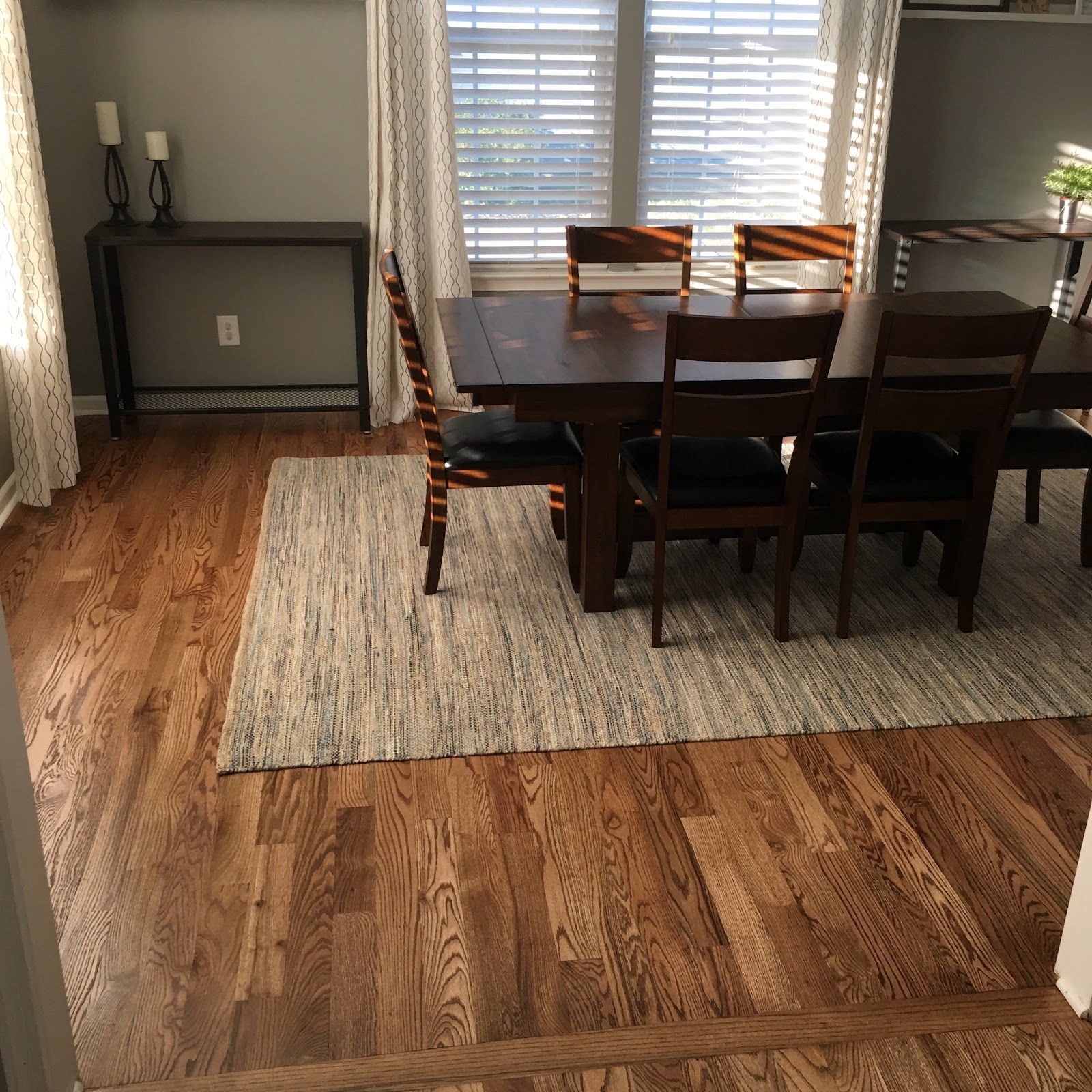 Dining room with dark wooden table, chairs, and rug on hardwood floor. Window with blinds and drapes.