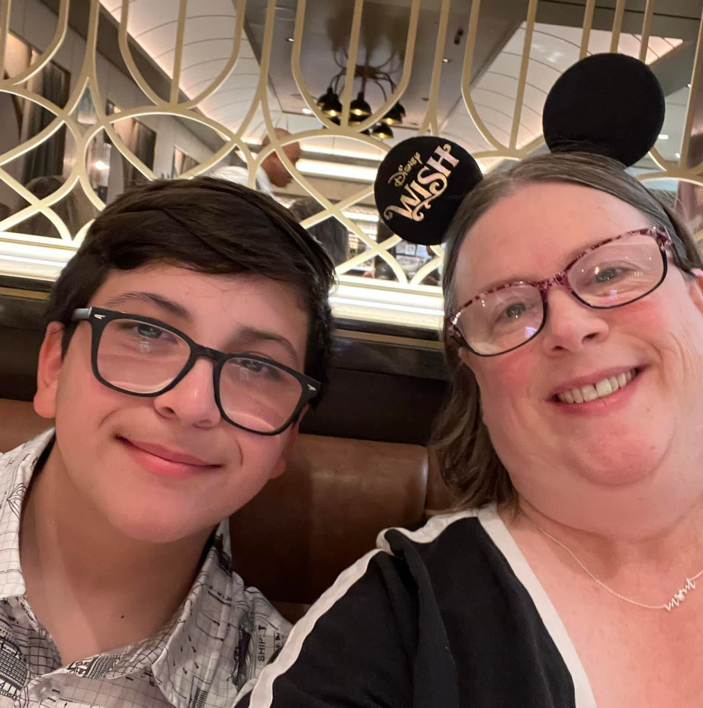 A person and a teenager smiling for a selfie wearing Disney Wish ears in a room with a decorative gold geometric wall.