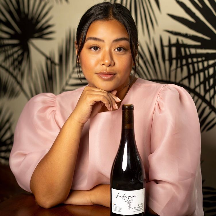 Woman in a pink blouse posing behind a wine bottle against a leafy background