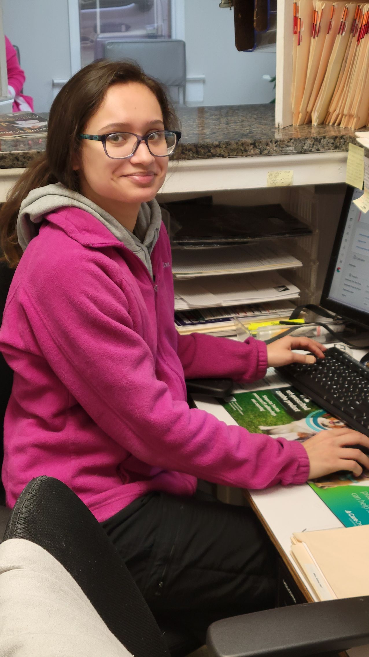 Person wearing glasses and pink hoodie, working at a desk with a computer, smiling.