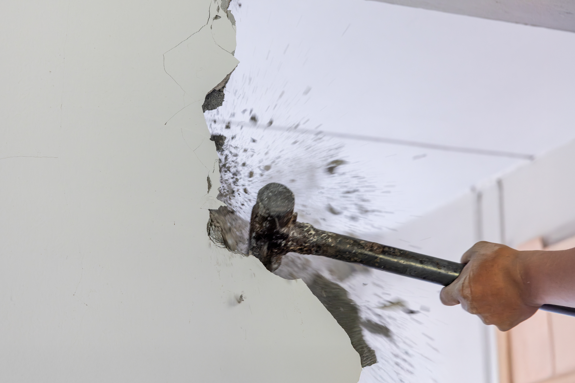 A person using a sledgehammer to break through a white wall, debris flying outwards.