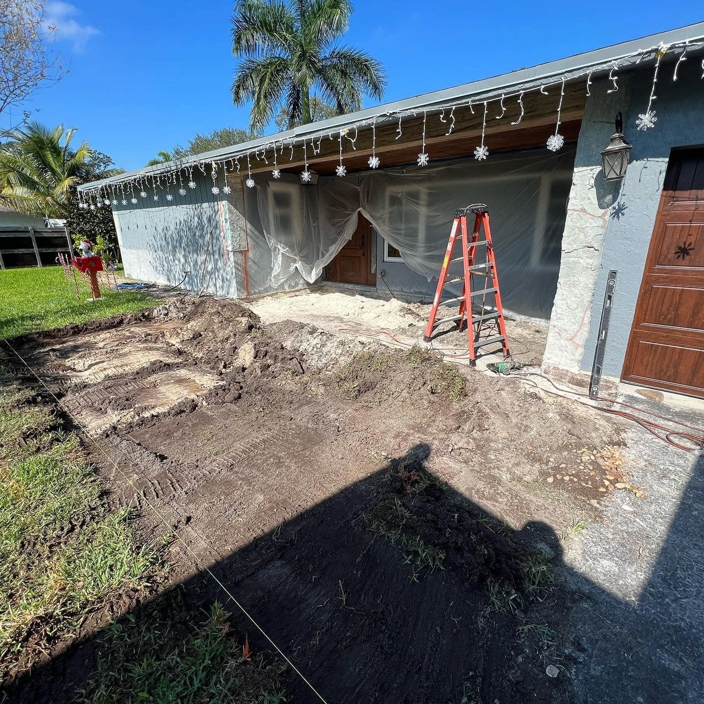 Exterior view of a house under construction; dirt patch in front, ladder, tarp over doorway, and blue sky.