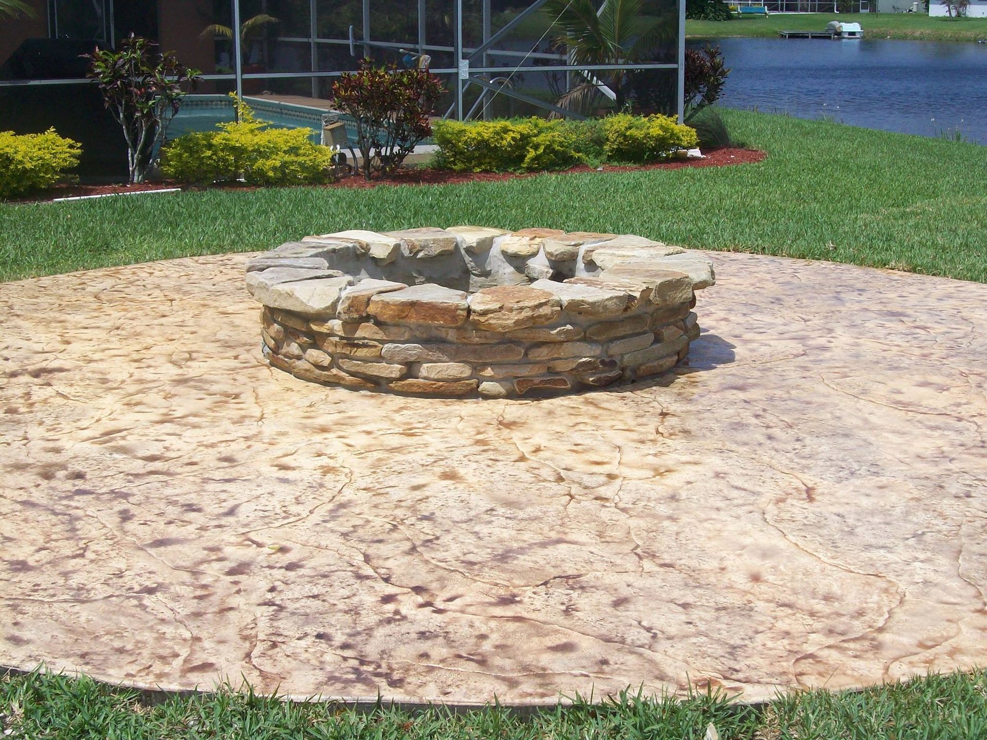 Stone fire pit on a textured concrete patio in a grassy backyard near a lake.