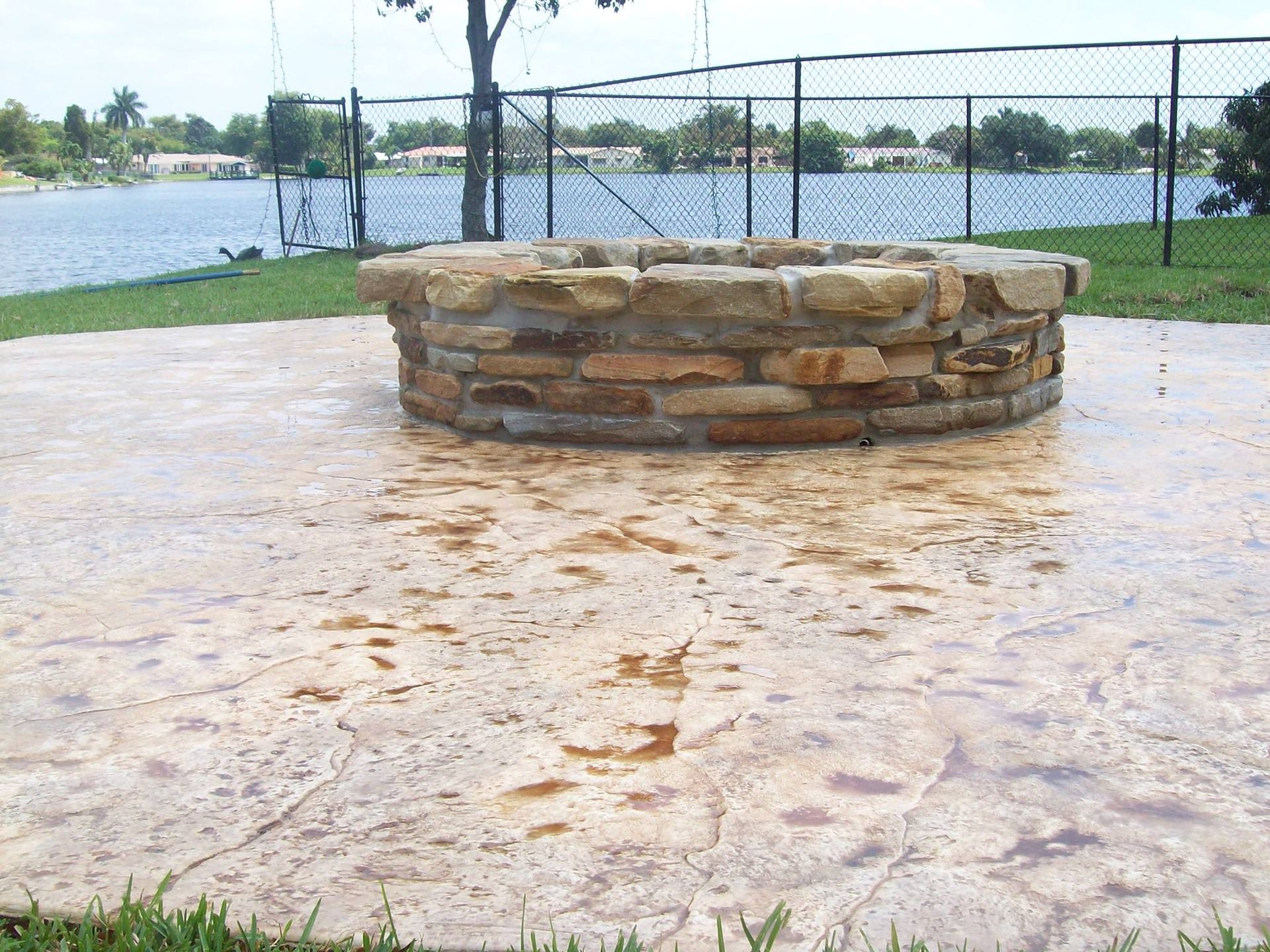 Stone fire pit on a textured concrete patio near a lake, green grass, and fence.
