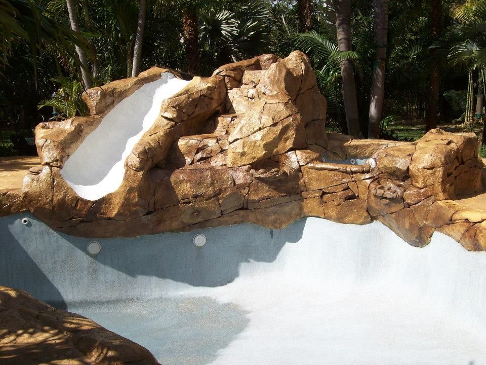 Empty pool with rock waterfall and slide feature, set in a tropical environment.