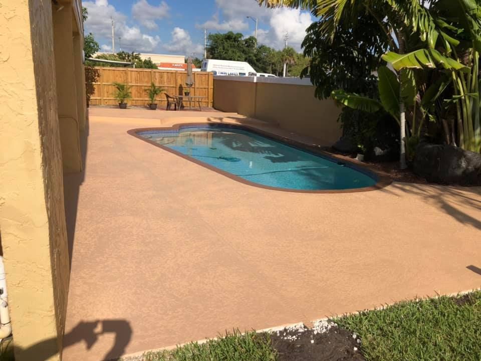 A backyard with a brown-colored pool deck and a turquoise pool. Green grass and bamboo fencing are also visible.