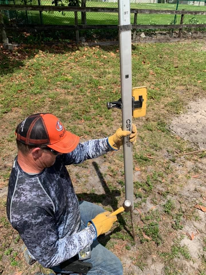 Man using a surveying rod with a level detector outdoors. He wears a hat, gloves, and is pointing at the rod.
