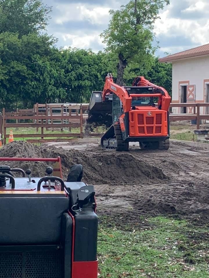 Orange Kubota skid steer moving dirt on a muddy construction site near a wooden fence and building.