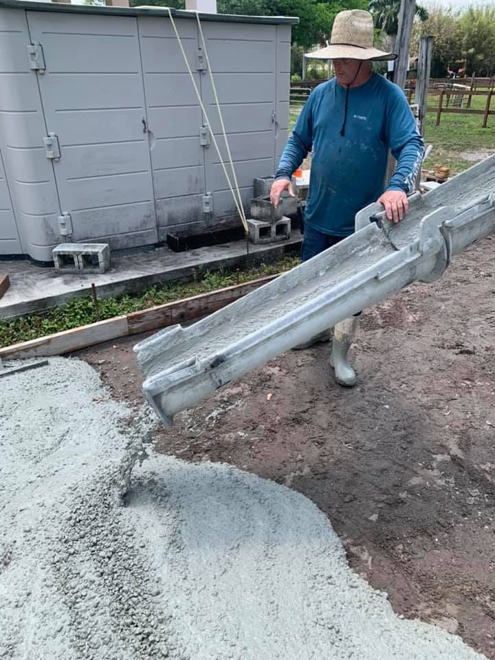 Man pouring concrete from a chute onto ground. He wears a hat and long-sleeved shirt. Concrete is gray; setting is outdoors.