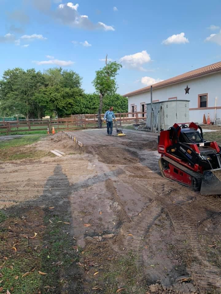 Construction site: Man stands on dirt path near mini excavator, white building in background.