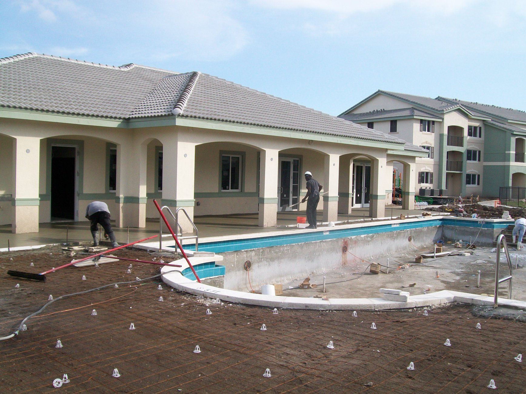 Pool construction near a tan house with workers. New construction with dirt and equipment.