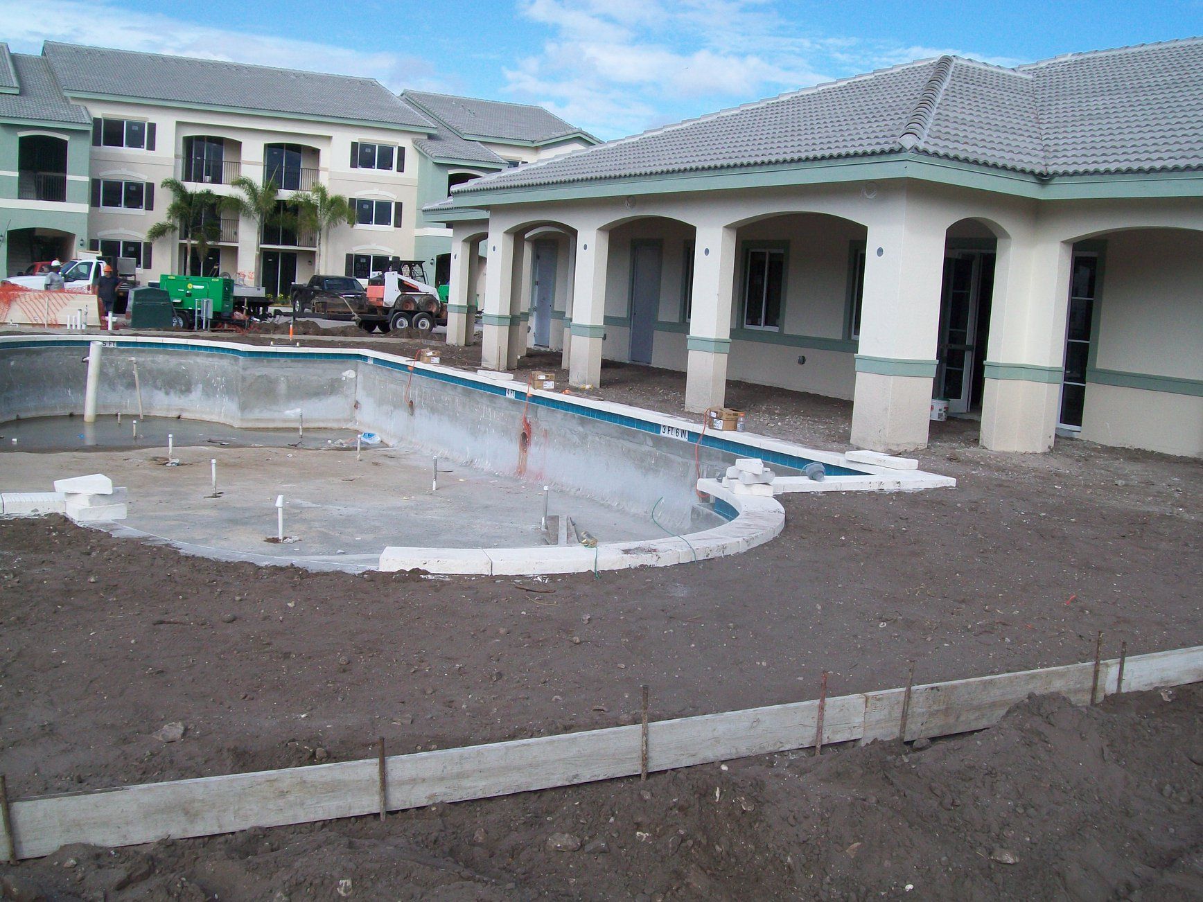 Pool under construction next to a building. Dirt and construction materials visible. Blue sky.