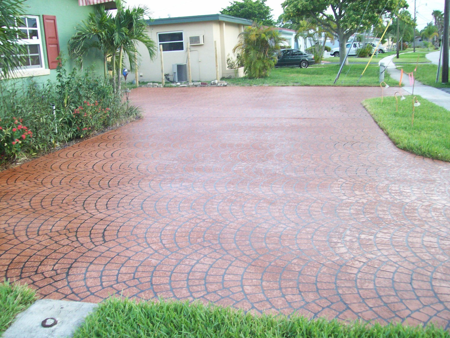 Red patterned concrete driveway in front of a green house with a small beige shed in the background.