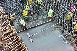 Construction worker inspecting concrete floor, taking notes with a measuring tool. Another worker in the background.