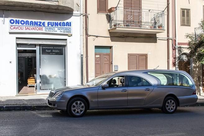 Carro funebre parcheggiato davanti a un'agenzia di pompe funebri italiana con un'insegna blu e bianca.