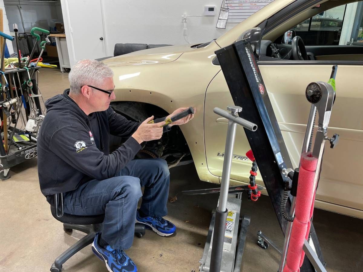 A person sits on a rolling stool, using a metal tool to perform paintless dent repair on the front fender of a beige car.