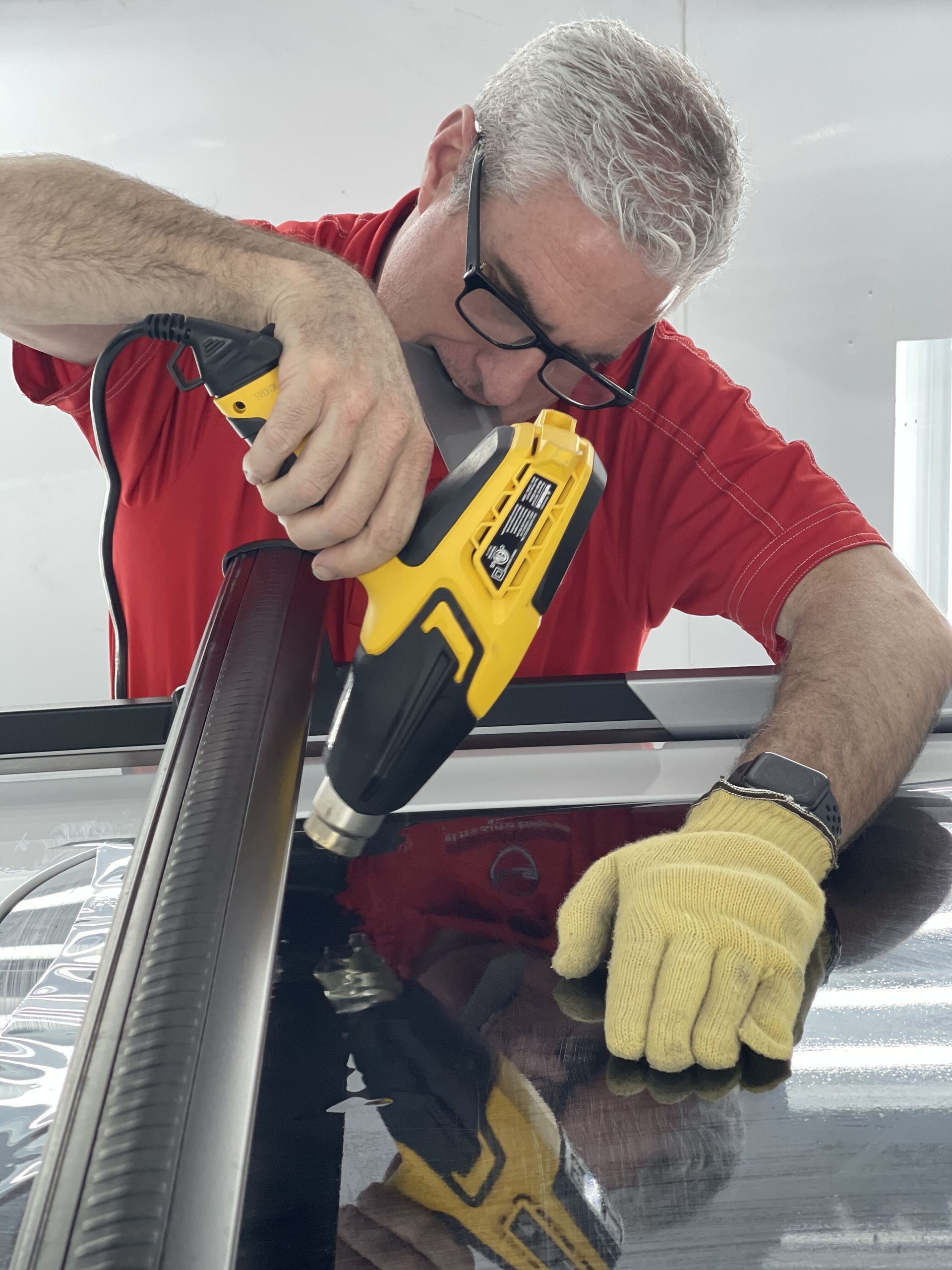 A person in a red shirt wears yellow gloves while using a heat gun to apply film to a vehicle surface.